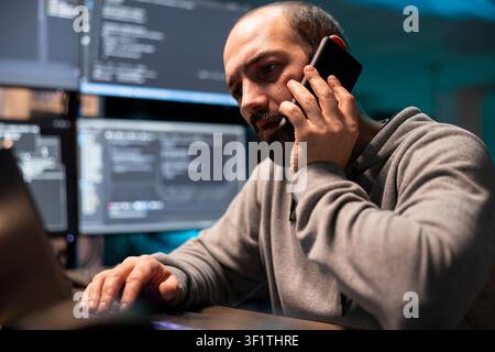 Cybersecurity specialist speaks on cellphone while typing at workstation, surrounded by multiple monitors displaying code. Programmer focused on addressing website issues and providing client support. Stock Photo