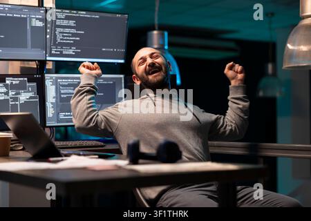 Male developer stretches back in excitement after completing a long coding task. Joyful programmer celebrates breakthrough moment in dim tech workspace filled with screens and digital tools. Stock Photo