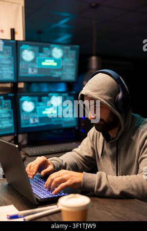 Bearded man in headphones concentrates on laptop as glowing monitors display technical interface. Freelance male programmer listens to music, working on software development codes in modern office. Stock Photo