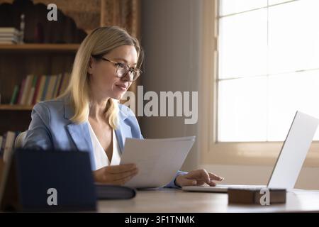 Older adult Caucasian businesswoman using laptop, reviewing printed documents Stock Photo