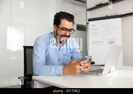 Smiling Arab businessman using smartphone at desk in corporate office Stock Photo