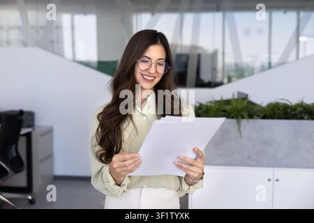 Smiling professional woman standing in corporate office reading documents Stock Photo