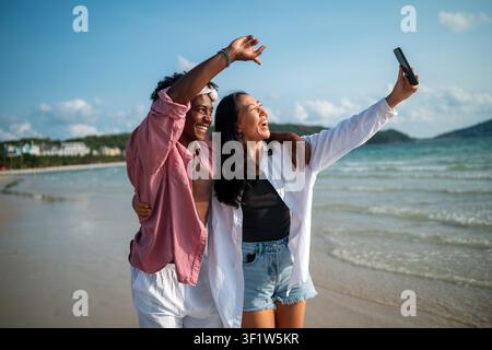 Trendy digital creator couple taking selfie by the beach, sharing travel content for social media Stock Photo