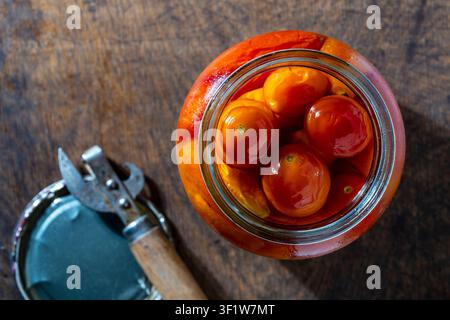 Canned red pickled cherry tomatoes inside an open glass jar on rustic wooden table with an old can opener, top view, close up. Flat lay food photograp Stock Photo