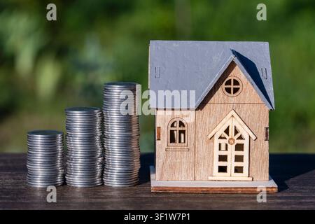 Wooden house model with growing stacks of coins. Miniature house model next to rising stacks of coins on a wooden table representing real estate inves Stock Photo