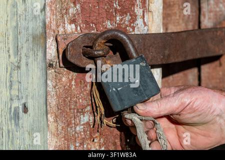 Male hand of an old man closes a rusty padlock on a wooden door, close up. Vintage background Stock Photo