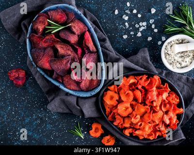 Baked beet and carrot slices with coarse sea salt scented rosemary. Vegan diet food idea and recipe. Healthy homemade beetroot and carrot chips in plate and ingredients on dark blue background. Copy space. Top view Stock Photo