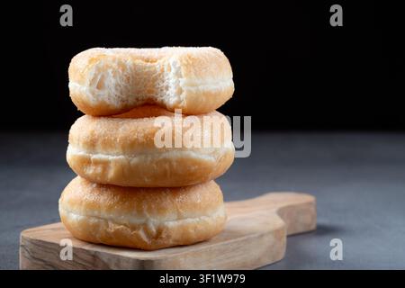 Vertical pile of golden fried sugar donuts on a wooden cutting board against a dark background, featuring a subtle bite on top Stock Photo