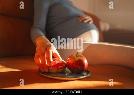 Pregnant woman resting on a sofa, reaching for a glazed donut from a small plate, with a fresh red apple sitting next to it, symbolizing food choices and cravings during pregnancy Stock Photo