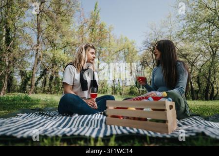 Two friends enjoying a picnic in a sunlit park sharing wine and conversation Stock Photo