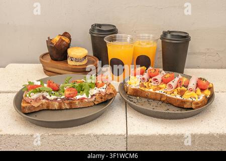 Two savory open-faced toasts with eggs, ham, and vegetables on gray plates, accompanied by orange juice, coffee cups, and pastries on a concrete ledge Stock Photo