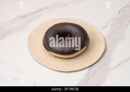 Chocolate glazed donut on a beige speckled plate over a white marble surface. Stock Photo