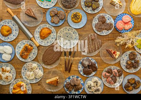 Overhead shot of various desserts like chocolate cookies, cake slices, and pastries arranged on blue patterned plates and glass dishes on a wooden tab Stock Photo