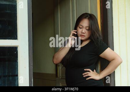 Asian woman standing by a doorway and talking seriously on her smartphone, wearing a basic black t-shirt. Indonesia. Stock Photo