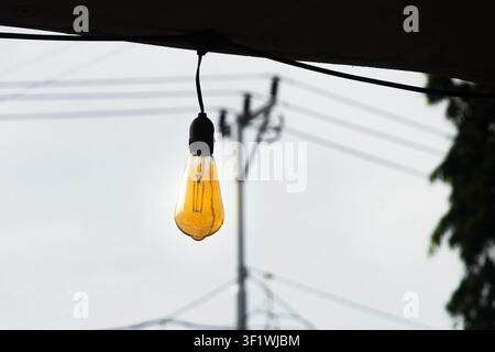 Close-up of a glowing vintage Edison-style light bulb hanging outdoors with blurred utility poles and power lines in the background Stock Photo