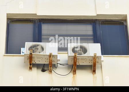 Two old air conditioning outdoor units with rusty metal brackets mounted on a building wall under a window. Stock Photo