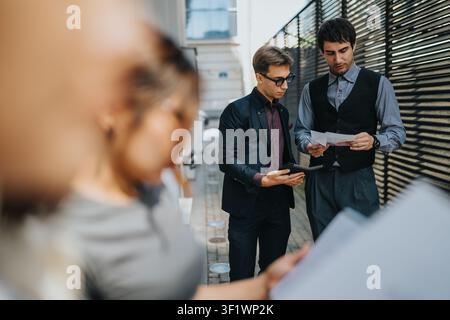 Two businessmen reviewing documents outdoors while a colleague reads notes in foreground Stock Photo