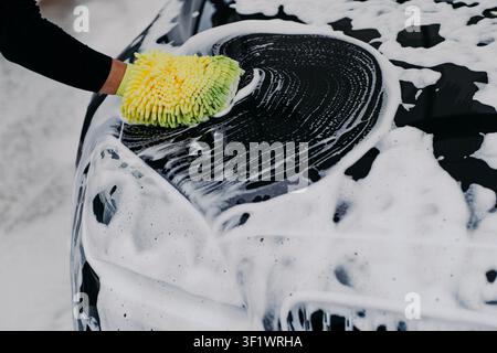 Person washes black car hood with yellow microfiber mitt and thick white foam at outdoor car wash in winter Stock Photo