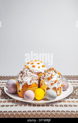 A collection of beautifully decorated Easter cakes and colorful eggs are presented on a white plate, set against a backdrop of a traditional embroidered cloth Stock Photo
