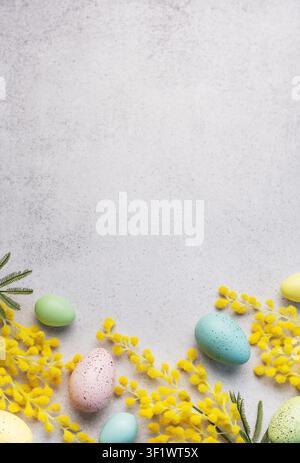 This image features a festive Easter arrangement of pastel-colored eggs and bright yellow mimosa flowers on a textured light grey surface. Stock Photo