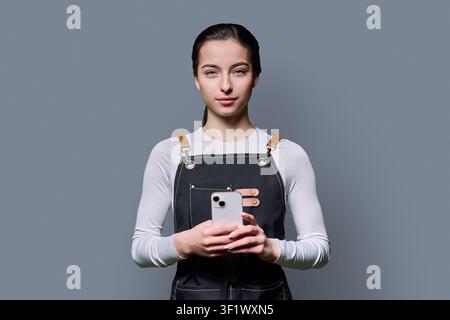 Confident young female in apron using smartphone posing on gray background Stock Photo