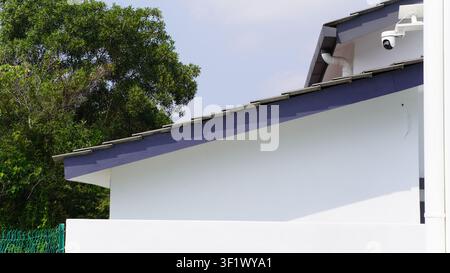 A pristine modern house exterior featuring a crisp white wall, sleek gray roof tiles, and a strategically mounted dome security camera under the eaves Stock Photo