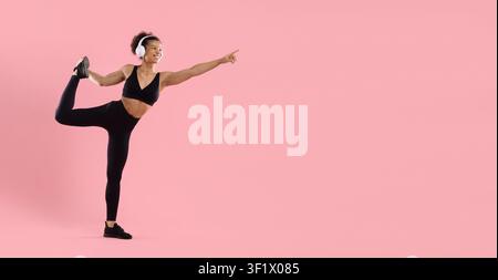 Flexible young Black fitness woman wearing headphones performing standing leg stretch Stock Photo
