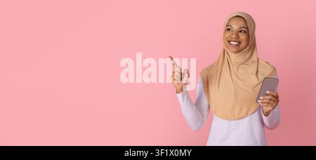 Black Muslim woman in hijab holding smartphone and pointing toward copy space Stock Photo