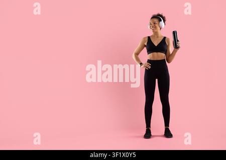 Black fitness woman wearing headphones holding reusable water bottle while looking aside Stock Photo