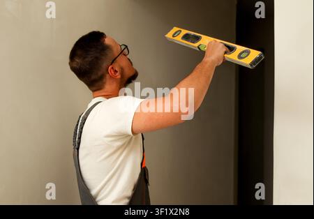 Professional builder in overalls checking wall alignment with spirit level. Construction worker or plasterer holding yellow level tool against concret Stock Photo