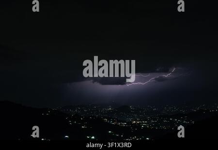 A dramatic night landscape showing a lightning strike above a mountain town in Himachal Pradesh with city lights shining against the dark sky. Stock Photo