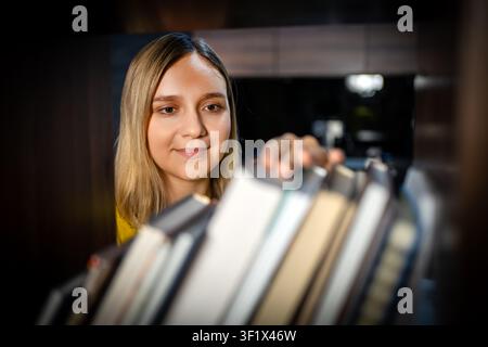 Woman choosing a book from a bookshelf, looking at the spines with a gentle smile. Bookstore or library, literary hobby, and reading selection. Stock Photo