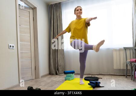 Tall young woman practicing yoga in a bright room with yoga mat. Focus on balance and strength during wellness session. Stock Photo