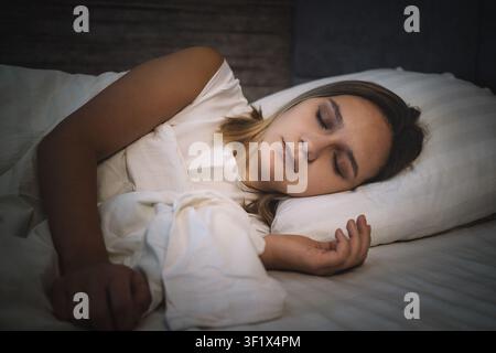 Woman sleeping peacefully on her side in a white bed with soft pillows. Deep sleep, restful night, and comfortable bedroom atmosphere. Stock Photo