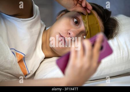 Morning routine and time management. Young sleepy woman in sleep mask checks her smartphone after waking up in bed, shocked by an alarm, unexpected me Stock Photo