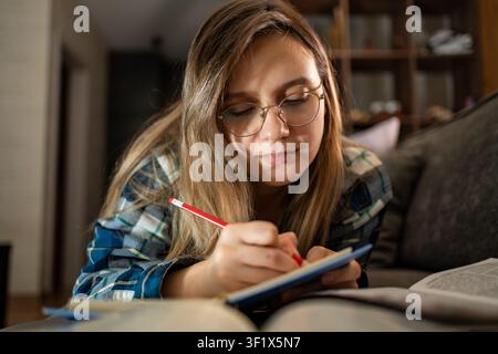 Smart student woman writing in a notebook while lying on a sofa with a stack of books. Home studying, creative writing, and cozy student lifestyle. wr Stock Photo