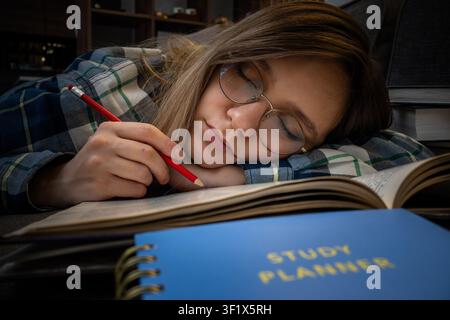Exhausted female student sleeping on an open book with a red pencil in her hand, next to a blue study planner. Academic burnout, late-night studying, Stock Photo