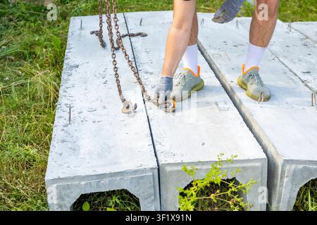 Worker removes lifting chains from precast concrete drainage channels on rural construction site. Drainage installation process Stock Photo