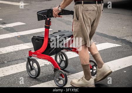 An elderly man pushes a red rollator across a pedestrian crossing in New York City. Stock Photo