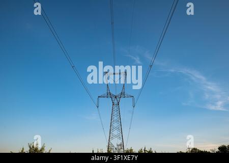 A low angle view of a tall metal transmission tower supporting overhead power lines extending across a bright blue sky with wispy clouds during the da Stock Photo