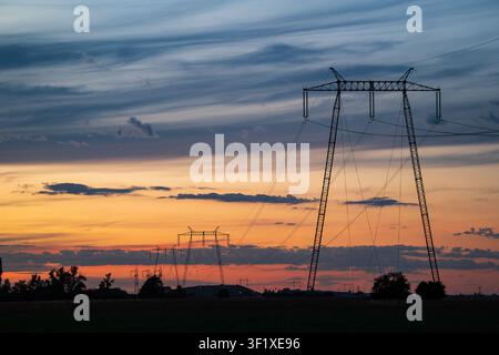 A wide shot showing the dark silhouette of massive steel utility pylons and power lines set against a vibrant orange and red sunset sky with dramatic Stock Photo