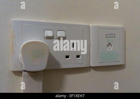White wall-mounted power outlets and a plug. Plug inserted into socket with visible green speckles. Outlets show standard UK design with two sockets Stock Photo