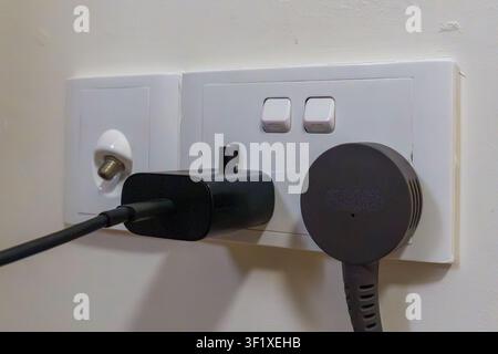 Black charger plugged into white wall socket. Plug and adapter attached to wall outlet. Simple, everyday electrical device scene. Minimalist design sh Stock Photo