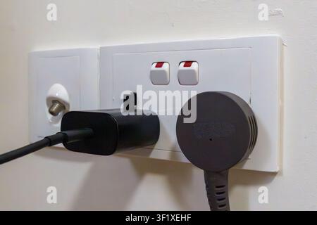 Black charger plugged into white wall outlet. Two red switches visible beside sockets. Cable extends from charger to unseen device. Plug design shows Stock Photo