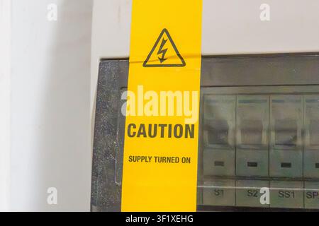 Yellow warning tape attached to electrical panel. Caution label shows active power supply. Warning symbol indicates high voltage risk. Panel includes Stock Photo