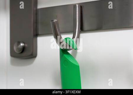 Green handle hangs from a metal hook. Hook mounted on a wall. Handle has a curved, sturdy design. Wall is plain and light-colored. Simple, functional Stock Photo