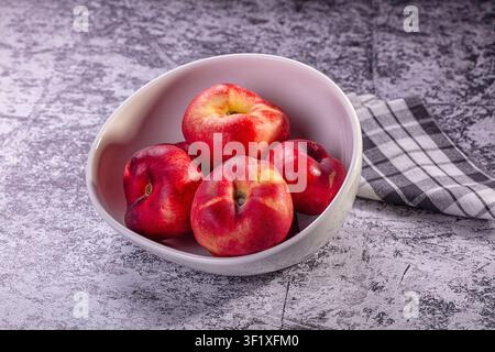 Ripe red nectarines arranged in white bowl on rustic gray surface. Soft natural lighting highlights glossy skin warm tones. Checkered cloth adds textu Stock Photo