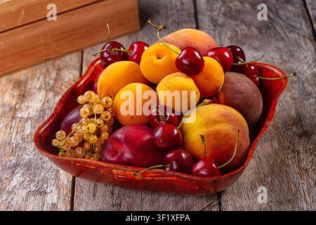 Vibrant apricots, cherries, peaches, nectarines, white currants arranged in red wooden bowl on weathered wood surface. Warm natural lighting highlight Stock Photo