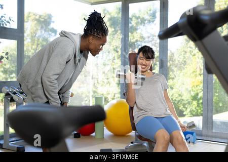 Male trainer leaning forward coaching female client doing bicep curl on gym bench with dumbbells Stock Photo