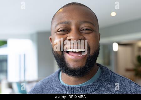 Mature African man wearing gray sweater laughing in living area near bright window and sofa cushion Stock Photo